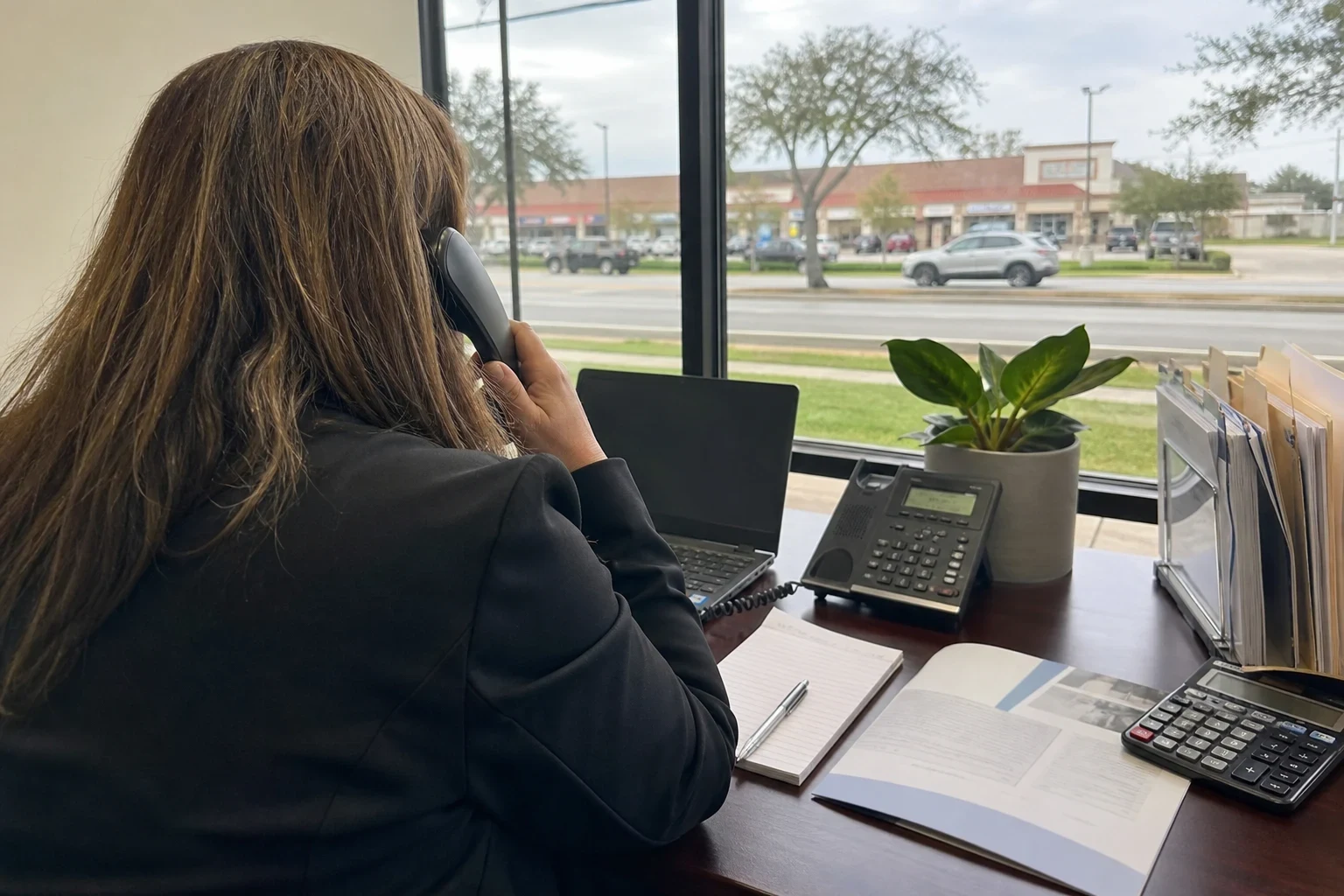 Agent seen from behind on a desk phone at an office desk with a notepad, booklet and calculator, window overlooking a road and strip center