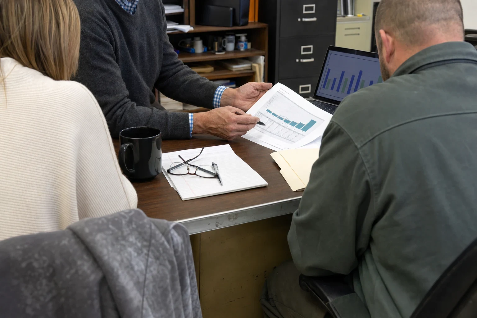 An agent points a pen at a printed bar-chart coverage illustration while a man and a woman sit across a worn metal desk in a back office with filing cabinets